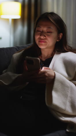 Vertical-Video-Of-Woman-Spending-Evening-At-Home-Lying-On-Sofa-With-Mobile-Phone-Scrolling-Through-Internet-Or-Social-Media